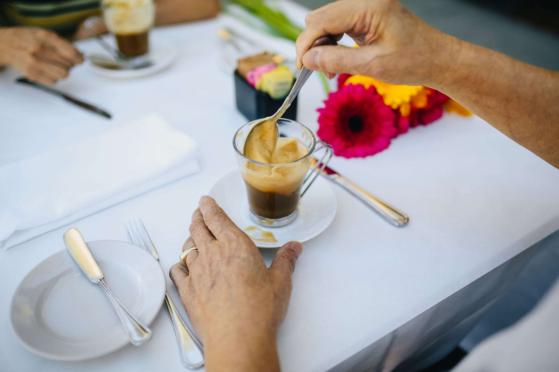 A person stirs coffee at a table set with flowers and cutlery, creating a cozy café scene.