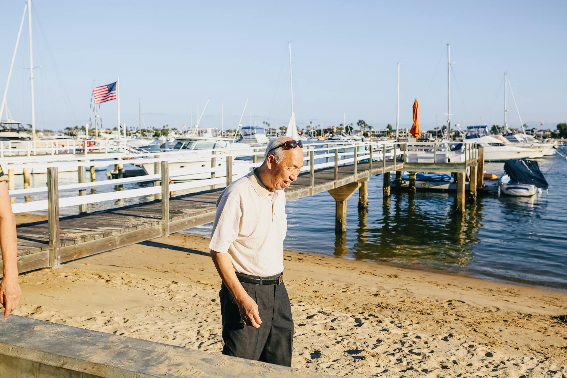 A senior man strolling by a marina with boats and a pier on a sunny day, enjoying leisure time.