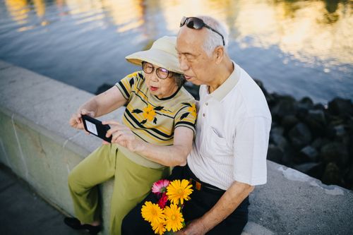 Free An elderly Asian couple takes a cheerful selfie by a waterfront while enjoying a sunny day. Stock Photo