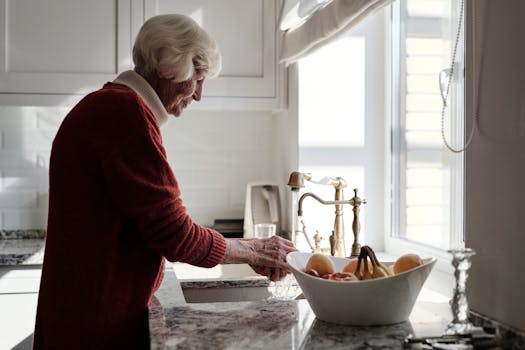 Elderly woman washing fruits at a kitchen sink bathed in sunlight.