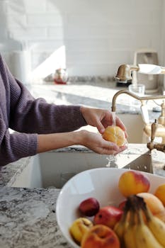 Close-up of hands washing a peach under running water in a sunny kitchen.