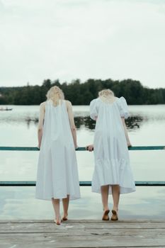 两名身穿白色连衣裙的女子赤脚站在湖边的码头上，凝视着平静的水面