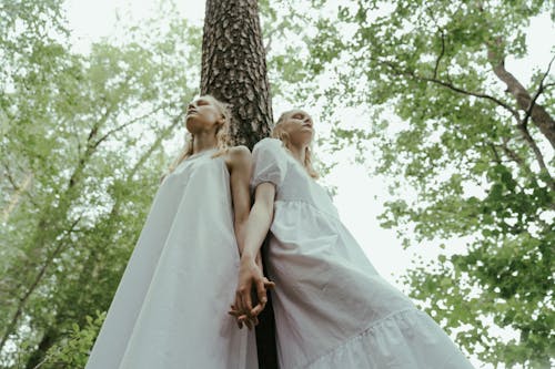 Free Low-Angle Shot of Twin Sisters Leaning on a Tree while Holding Hands Stock Photo