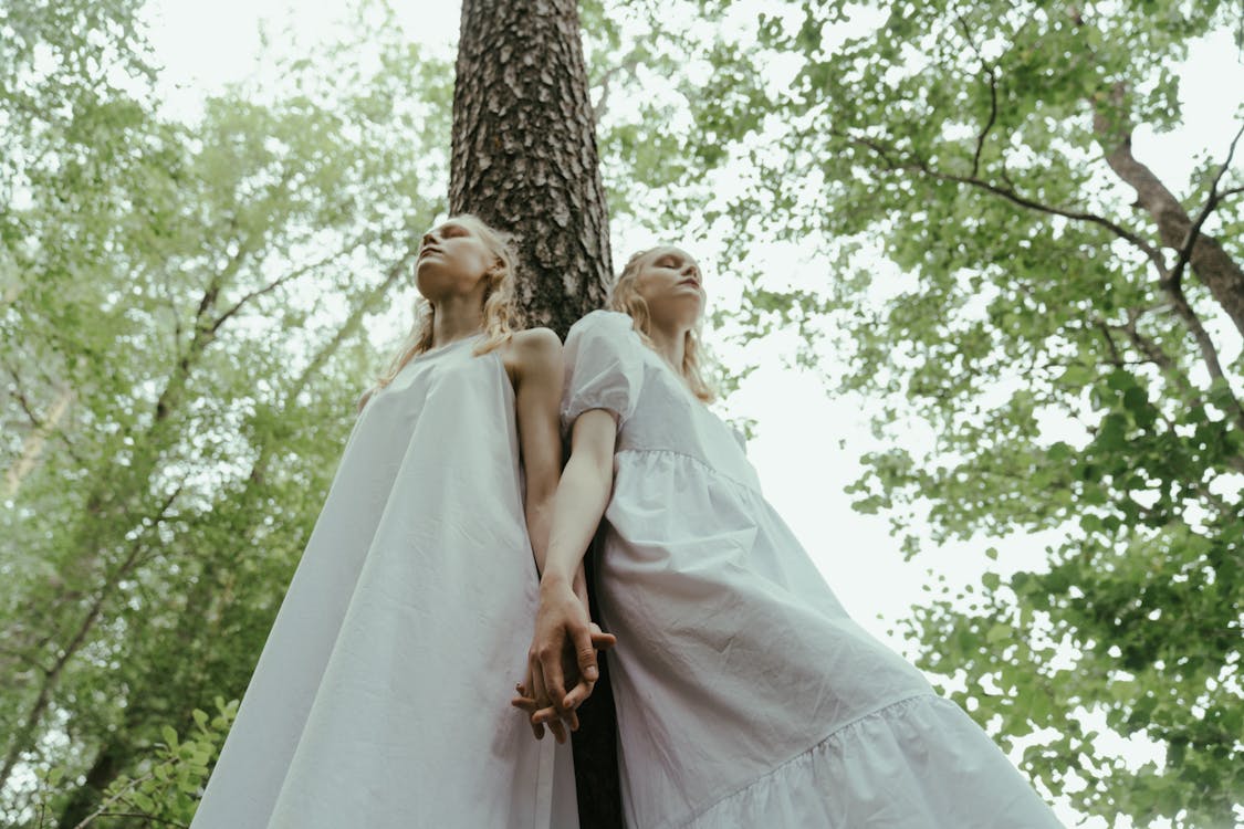 Low-Angle Shot of Twin Sisters Leaning on a Tree while Holding Hands