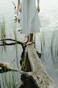 一位赤脚女子身着白色连衣裙，优雅地站在平静湖边的一根木头上，四周环绕着大自然