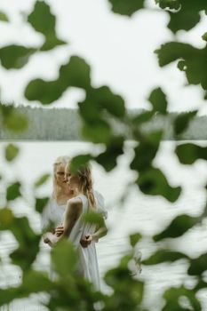 两个穿着白色连衣裙的女人在宁静的湖边拥抱，身后映衬着树叶