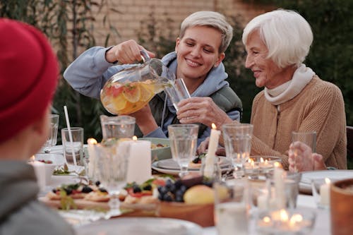 Positive lady pouring drink into glass for elderly woman while having dinner with family on terrace