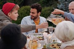 Faceless female pouring fruit beverage into glass of boy in warm cloth while cheerful multiethnic family having dinner outdoors