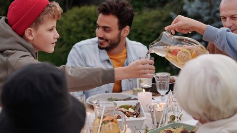 Faceless female pouring fruit beverage into glass of boy in warm cloth while cheerful multiethnic family having dinner outdoors