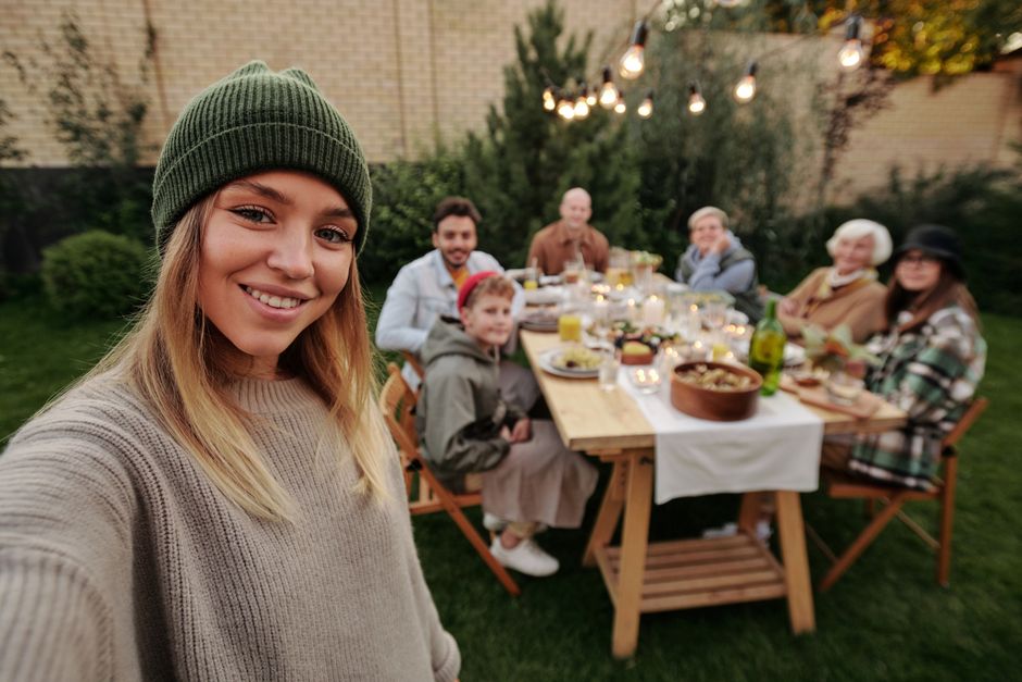 Photo by Askar Abayev on Pexels A joyful family dinner outdoors, captured with a smiling woman in the foreground taking a selfie.