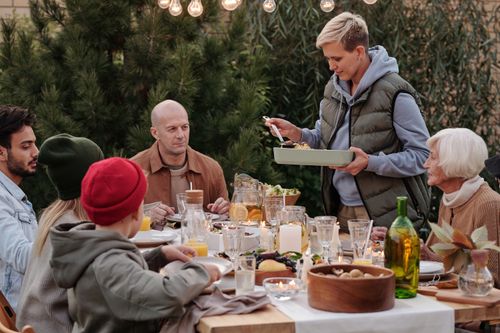 Free Smiling woman wearing warm vest and hoodie serving baked dish during holiday party at big table under garlands and evergreen trees beside Stock Photo