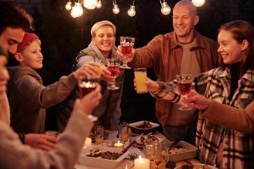 Laughing friends with kids spending time in backyard at night enjoying dinner with garlands and clinking glasses