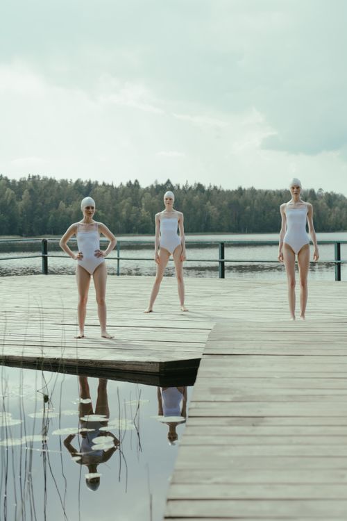 Free Three models in swimsuits pose on a wooden pier by a tranquil lake, emphasizing summer fashion. Stock Photo