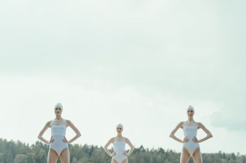 Free Three models in white swimsuits pose outdoors with a serene sky background, embodying elegance and symmetry. Stock Photo