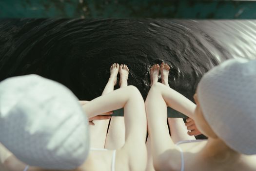 两名妇女戴着泳帽、穿着泳衣坐在水边，把脚浸入水中