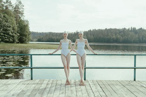 Two Women in White Swimsuit Standing Side by Side on a Wooden Dock