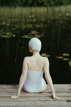 一位穿着泳衣、戴着帽子的女人，正坐在宁静湖边的码头上，四周环绕着大自然