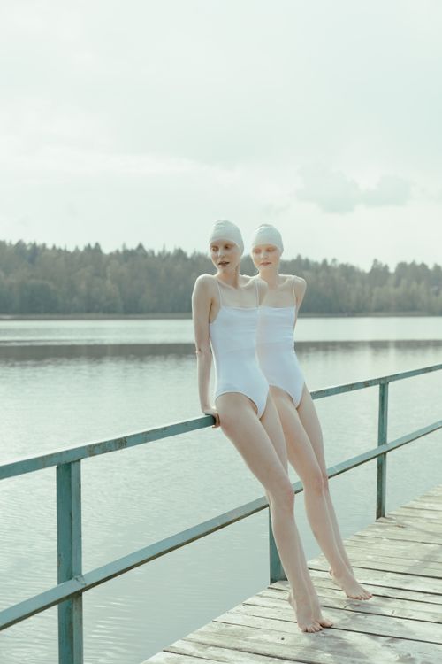 Free Two women in swimsuits lean against a pier by a scenic lake with forest backdrop. Stock Photo