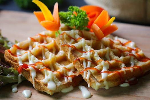 Close-up of crispy waffles drizzled with sauces, garnished with fresh herbs and vegetables on a wooden board.