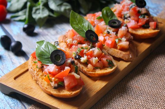 Toasted bread topped with diced tomato, basil leaves and black olive rings on a wooden board