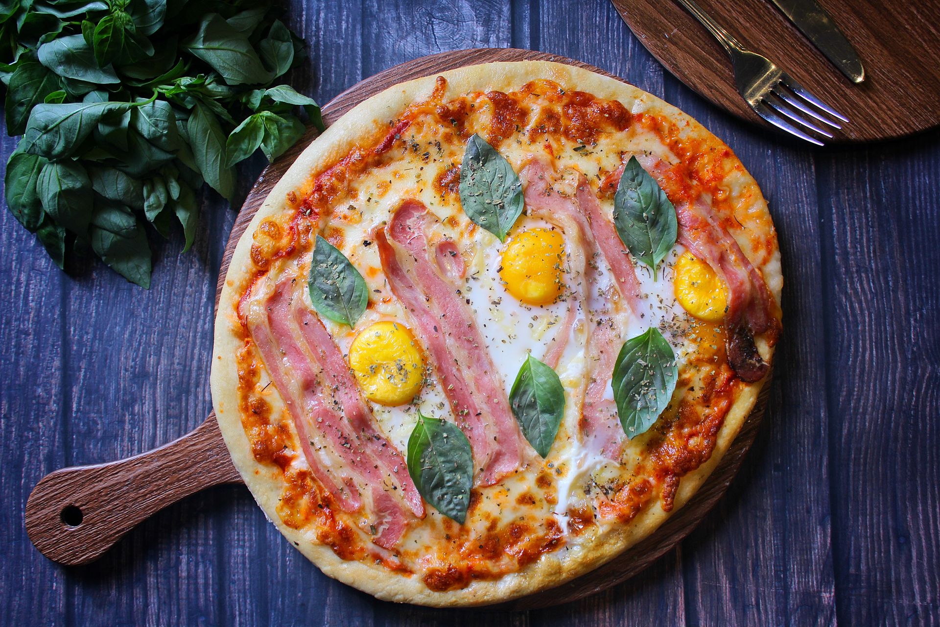Delicious pizza topped with ham, basil leaves, and egg yolks on a rustic wooden tray.