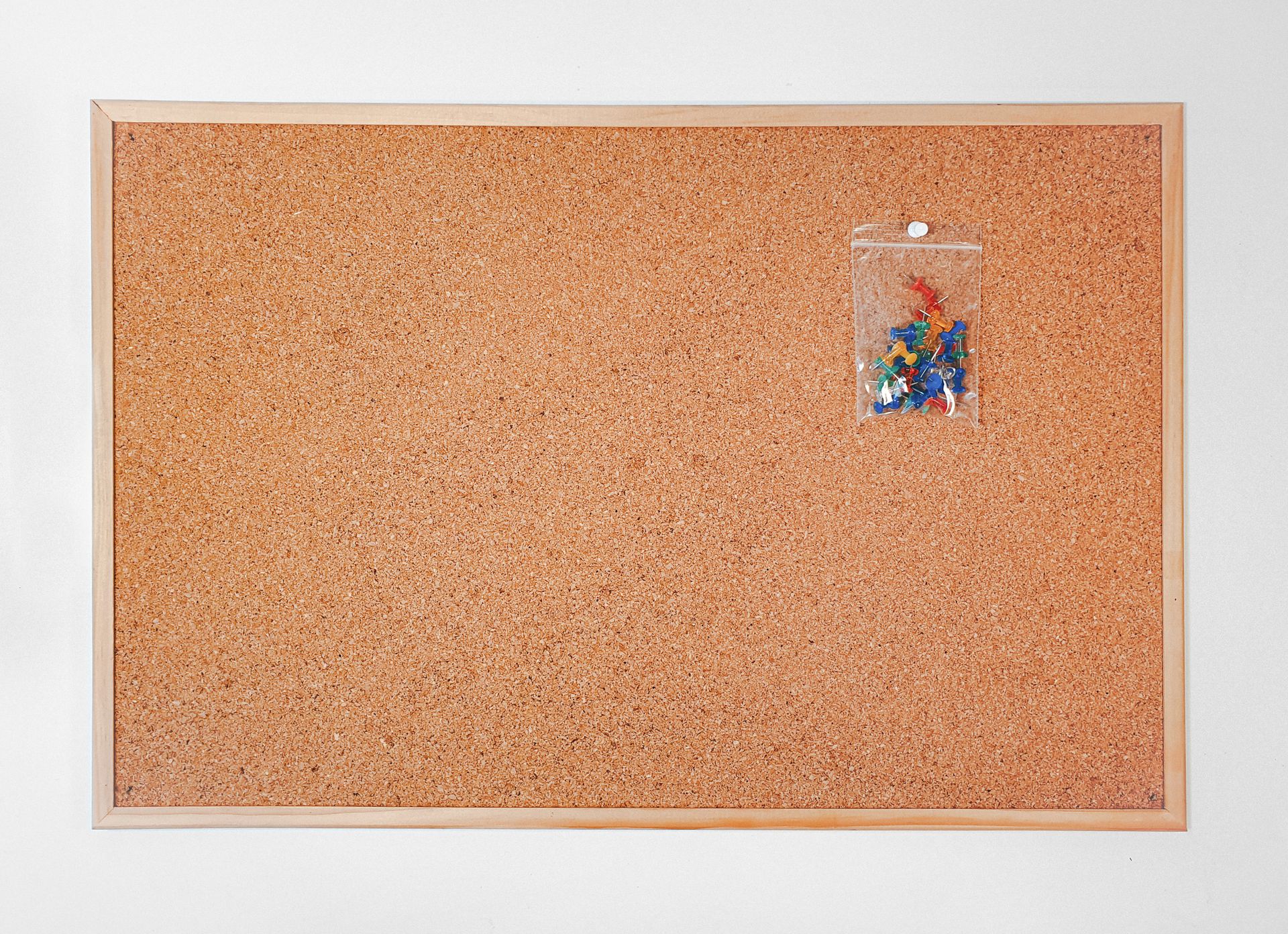 Cork board with wooden frame featuring a packet of colorful push pins for office or home use.