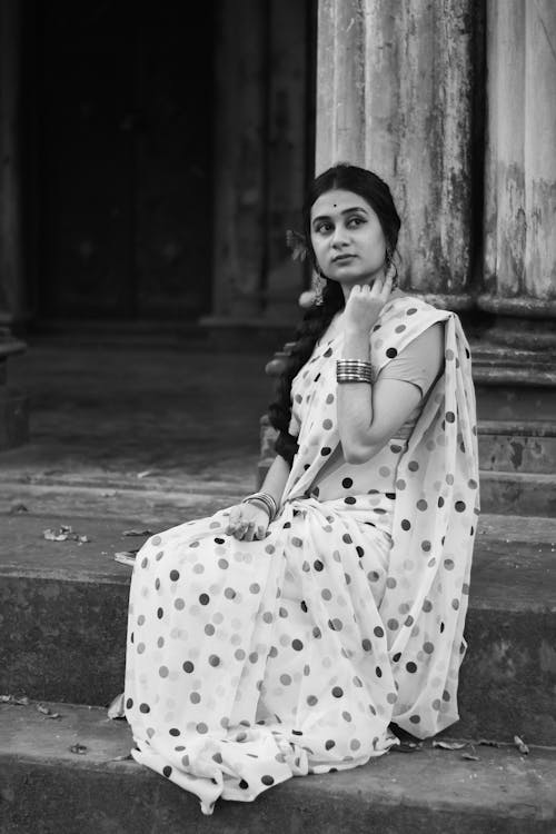 Black and white of charming young female wearing traditional sari sitting on stairs of old building and looking away