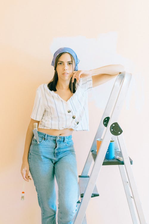 Woman in Striped Shirt and Denim Jeans Leaning against a Ladder near the Wall