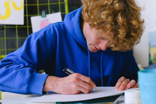 Free Young guy in casual outfit sitting at table in light workshop while cutting object with utility knife on paper near painting tools Stock Photo