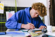Concentrated young guy in casual outfit sitting at table in light studio while cutting object from paper drawing with utility knife near paints and drawing tools