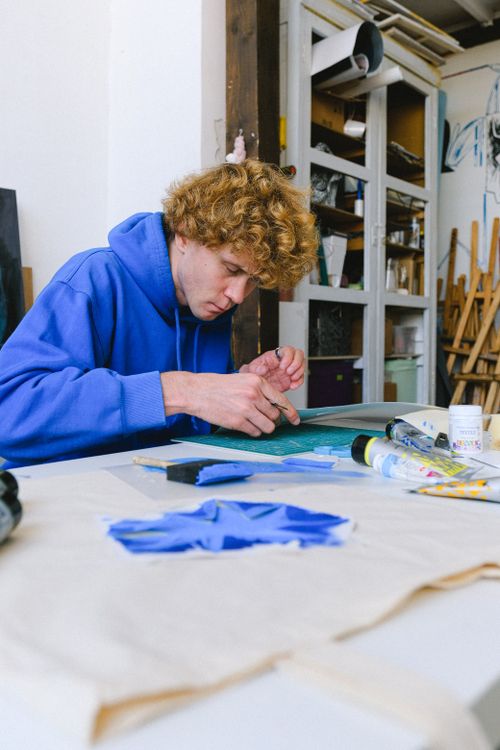 Free Young man in casual clothes sitting at table with stencil on paper creating drawing on shopping bag near paints and painting tools in light workshop Stock Photo