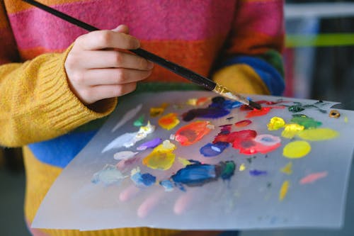 Crop anonymous female artist in casual clothes standing in light studio with paintbrush while mixing various paints on paper