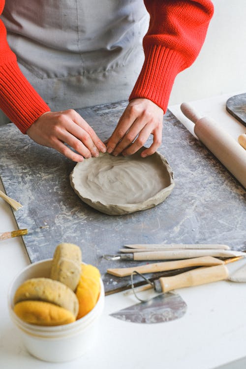 Crop faceless lady in apron standing near table while shaping piece of clay with hands near sponges and double end loops with stacks and rolling pin for pottery in studio