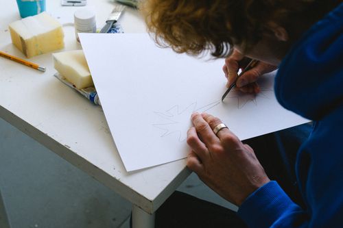 Free High angle of crop unrecognizable man in casual clothes sitting at table while cutting sketch on paper with utility knife near paints and sponge with pencil in light workshop Stock Photo