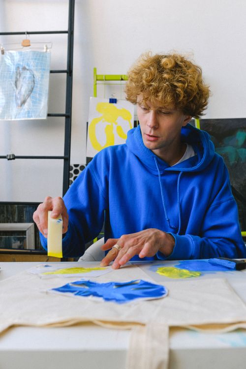 Free Concentrated young guy in casual outfit sitting at table in light studio while painting shopping bag with colorful elements and using sponge for creating artwork Stock Photo