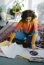High angle full length of young thoughtful woman in casual clothes sitting on carpet on floor while looking through paintings near head sculpture and potted plant in daylight in room