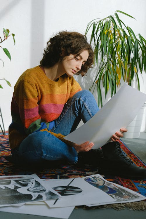 Free Young female artist sitting on floor with legs crossed and watching drawings in room Stock Photo