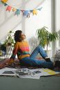 Side view of young female designer in smart casual outfit sitting on floor with drawings and thinking about new creative project in room