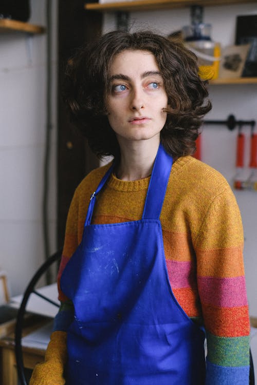 Young female artist in casual wear and blue apron thinking and looking away in arts craft shop