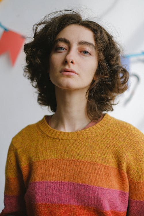 Low angle of young pensive curly haired female in colourful warm jumper looking at camera