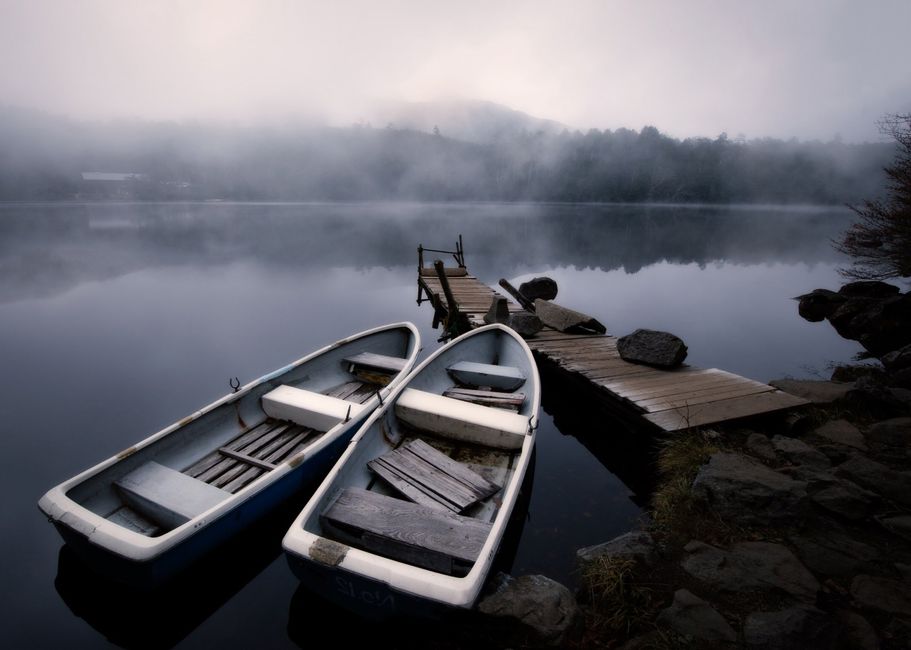 Old wooden pier near stone shore and moored boats on calm lake surface in mist