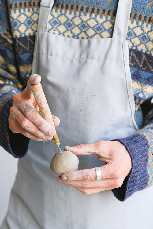 Unrecognizable male potter wearing apron standing with piece of clay in hand and sharp tool while  working in professional studio
