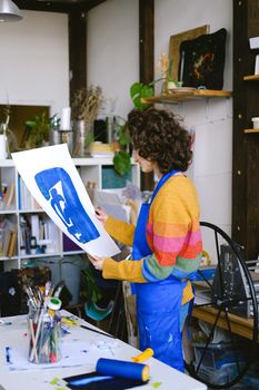 一位穿着五颜六色毛衣的女艺术家，在充满活力的工作室里拿着一幅蓝色的版画