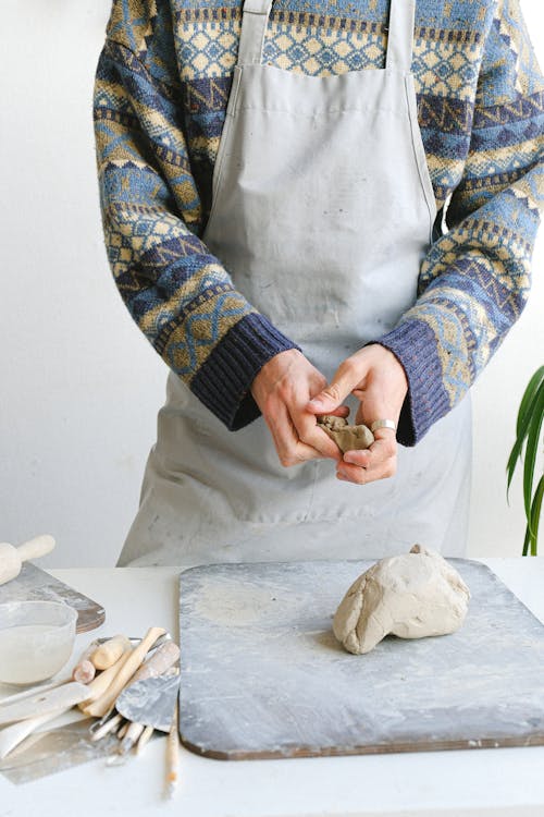 Unrecognizable male master in apron standing at table with piece of clay on wooden board while creating pottery in workshop