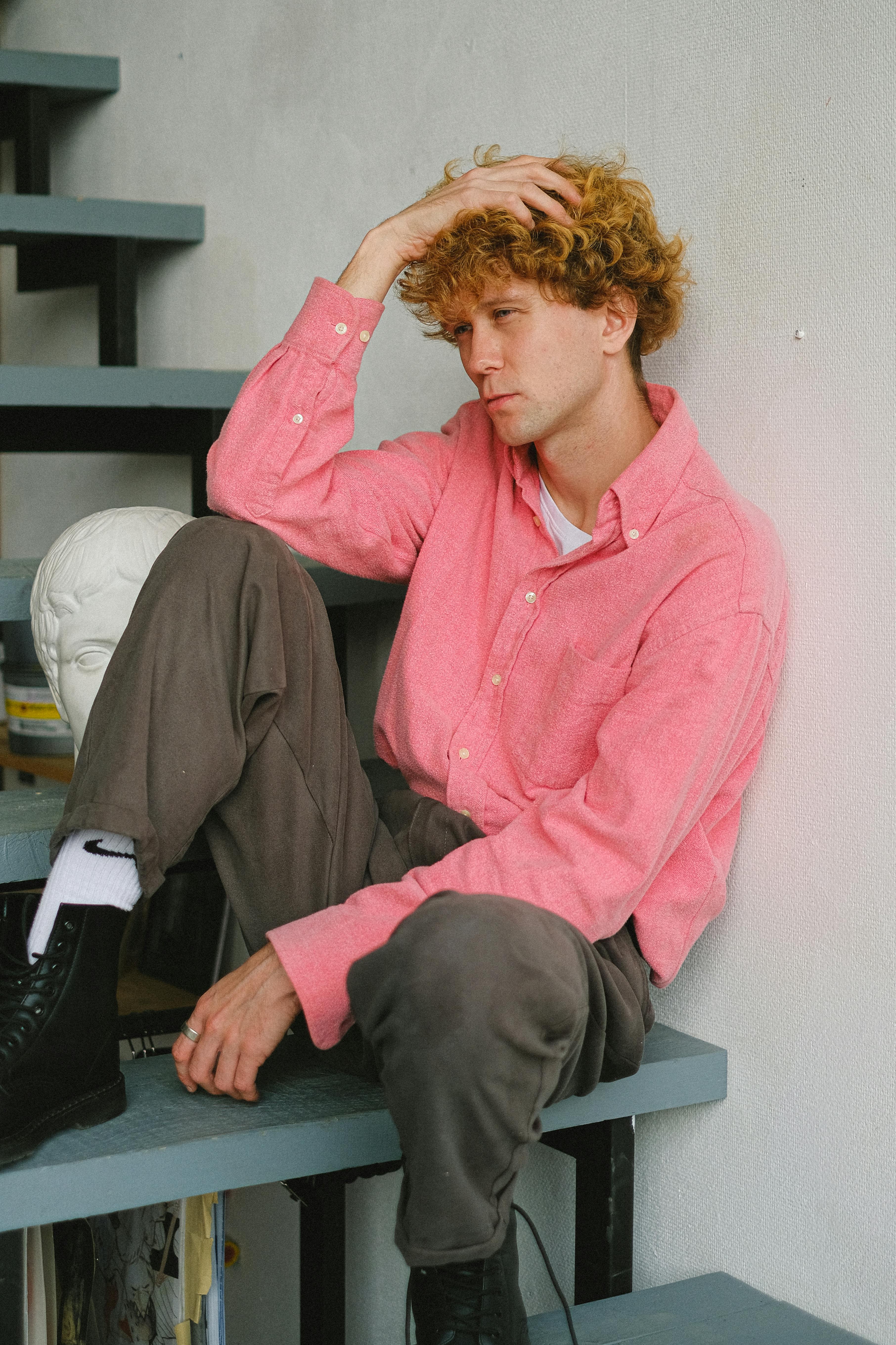 Side view of pensive male wearing pink shirt sitting on steps with hand on head near wall with historic bust