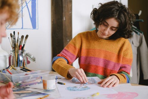 Free Concentrated female painter drawing sketches on paper with crayons while sitting at table near anonymous coworker in creative workshop during work Stock Photo