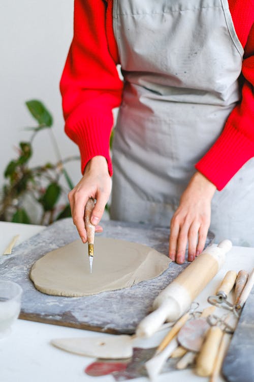 Unrecognizable female master with sharp tool cutting clay on wooden board while standing at table with various instruments in workshop