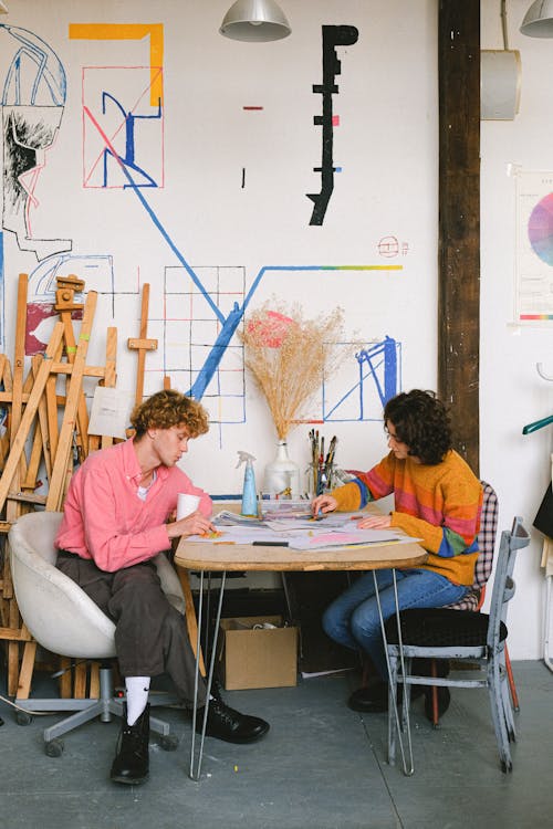 Free Full body side view of focused artists drawing pictures while sitting at table with art supplies in creative workshop with easels Stock Photo