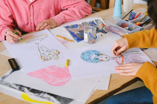 From above of talented artists drawing pictures with colorful crayons while sitting at table and creating paintings in art studio
