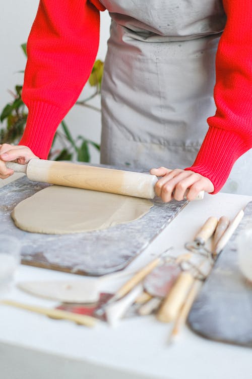 Unrecognizable female master standing at table with wooden rolling pin wearing apron rolling piece of clay while making pottery in professional workshop